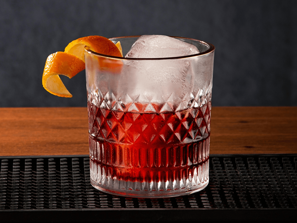 Negroni served over a large ice cube in a patterned rocks glass with an orange peel garnish.