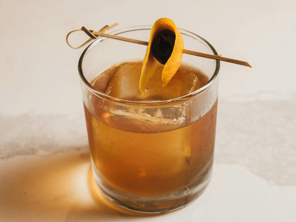 Amber cocktail served over a large ice cube with an orange peel and dark cherry garnish.