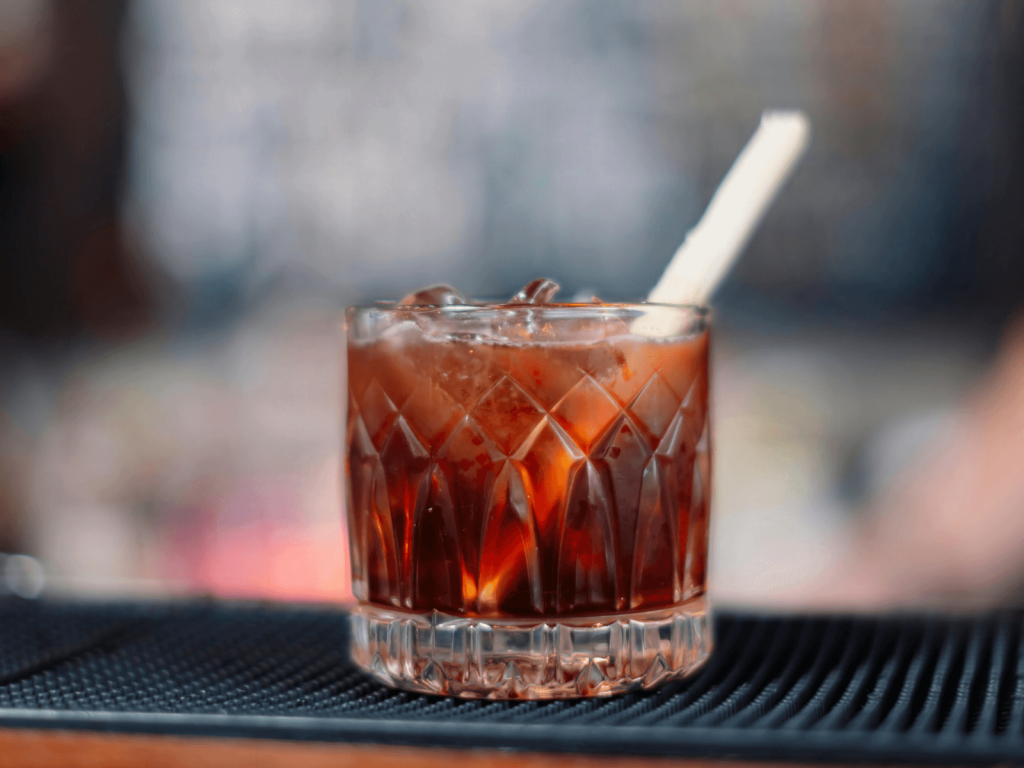 Dark red cocktail served over ice in a cut crystal rocks glass on a bar mat, with a blurred bar background,