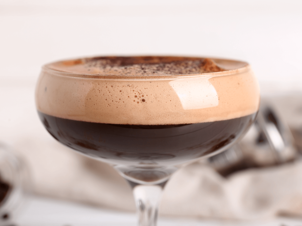 Close up of a dark espresso martini in a coupe glass with a thick layer of tan foam on top.