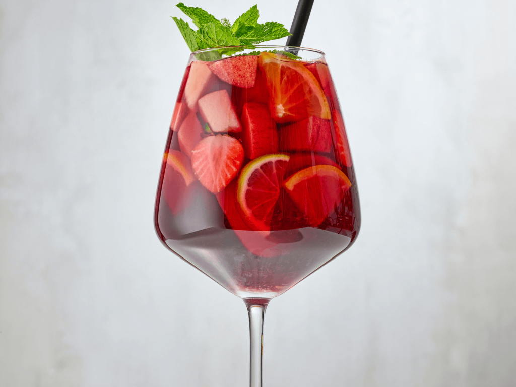 Red sangria in a large wine glass with sliced strawberries, citrus wheels, fresh mint, and a black straw against a light background.
