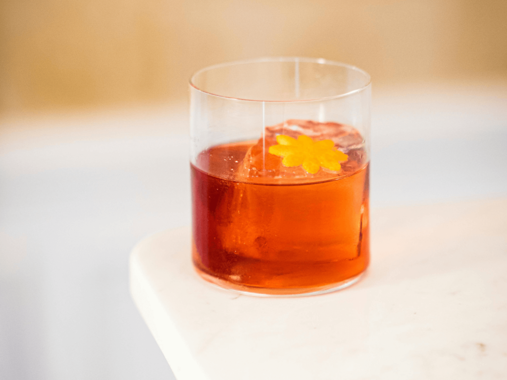 Negroni cocktail in a rocks glass with a large ice cube and a small yellow flower garnish on top.