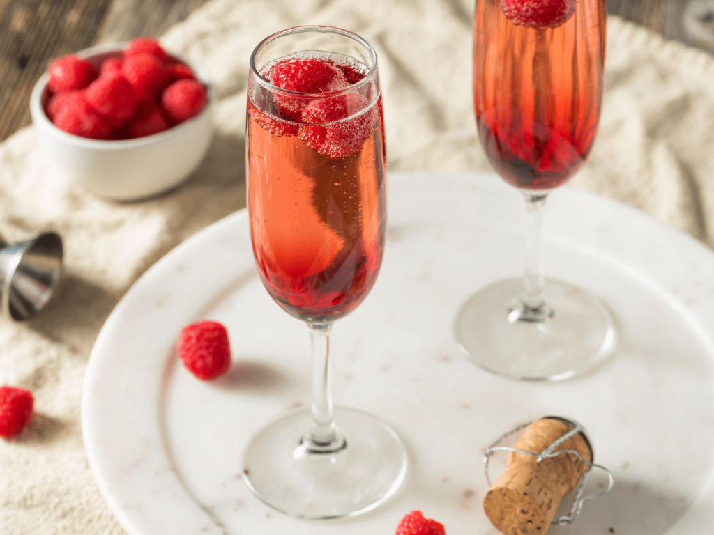 wo tall champagne flutes filled with pink sparkling cocktail, each garnished with raspberries, set on a white tray with a cork and fresh raspberries nearby.