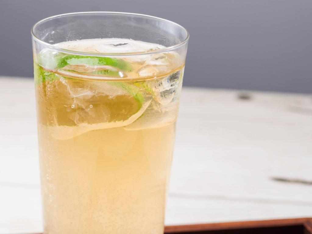 Tall highball glass filled with a pale, bubbly drink over ice, garnished with a lime wedge, representing international cocktails served simply.