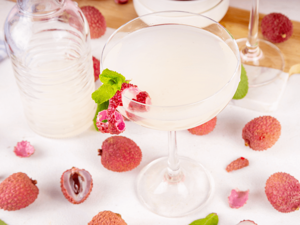 Pale lychee martini in a coupe glass with a mint leaf and a lychee garnish, surrounded by whole and halved lychees.