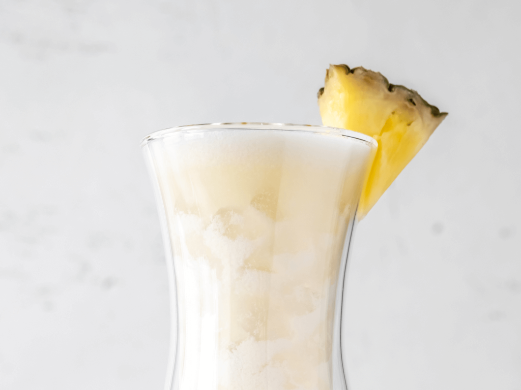 Creamy frozen piña colada in a tall curved glass with a pineapple wedge garnish, shown as one of the world cocktail recipes you can make at home.