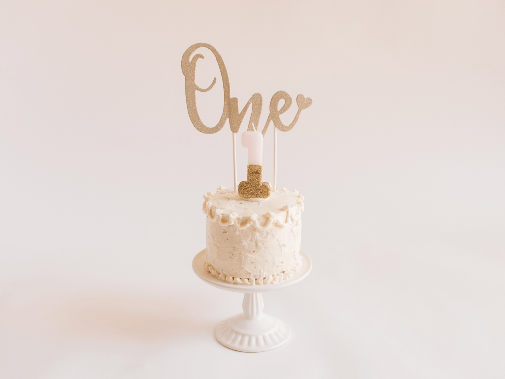 White first birthday cake on a cake stand with a gold “One” topper and pink number one candle, perfect for a post about 1st birthday wishes and card messages.