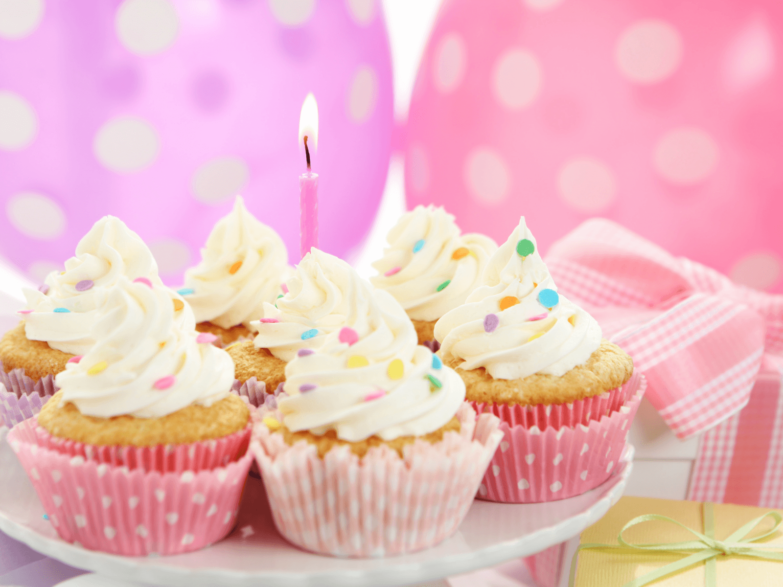Cupcakes with white frosting, colorful sprinkles, and a lit pink candle in front of pink balloons for a baby’s first birthday celebration.