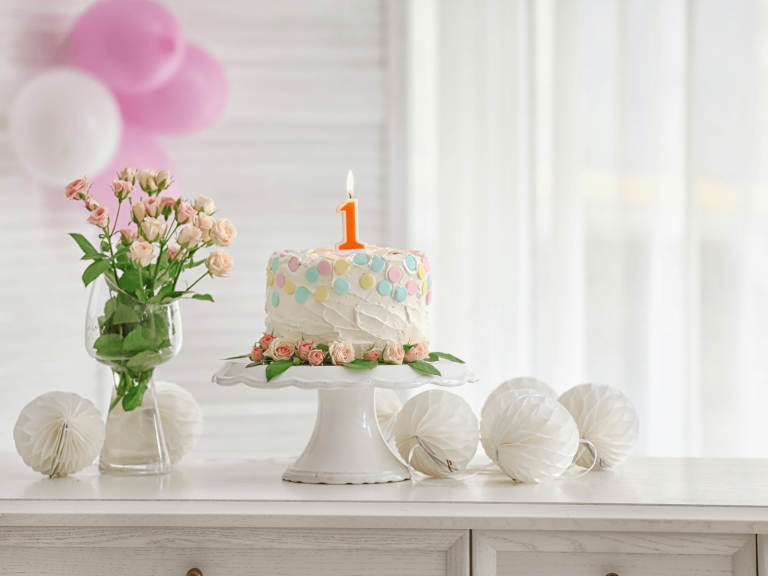 White first birthday cake with a lit number one candle, pastel decorations, pink balloons, and flowers, used for a post about 1st birthday wishes for cards and captions.