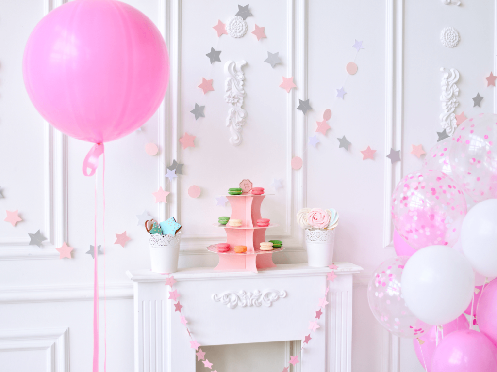 Pink and white first birthday dessert table with balloons, star garland, macarons, cookies, and flowers for a baby’s first birthday celebration.