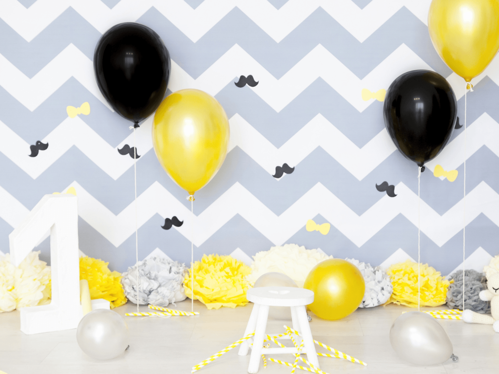 First birthday party setup with black and yellow balloons, a large white number one, a small white stool, and a gray chevron backdrop.