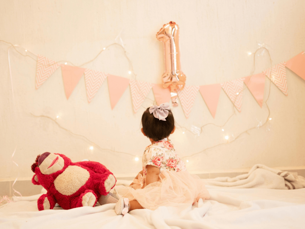 Baby girl sitting on a bed beside a stuffed bear with pink party decorations and a number one balloon, celebrating her first birthday.