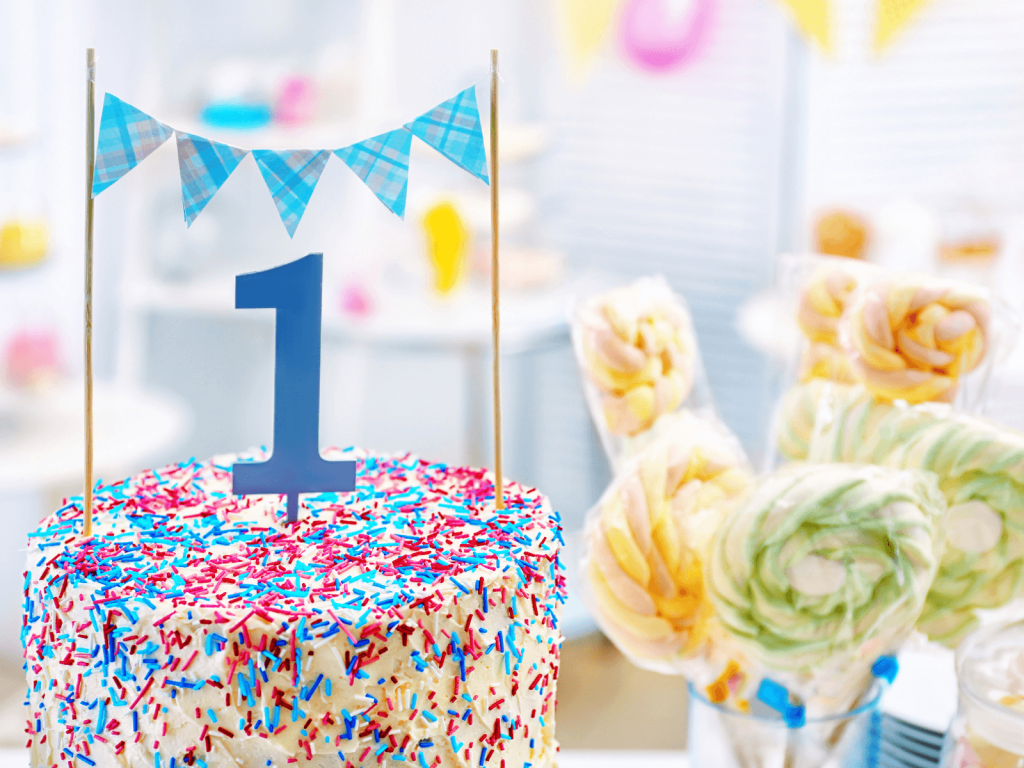 Close-up of a first birthday cake with colorful sprinkles, a blue number one topper, and a small blue bunting decoration.