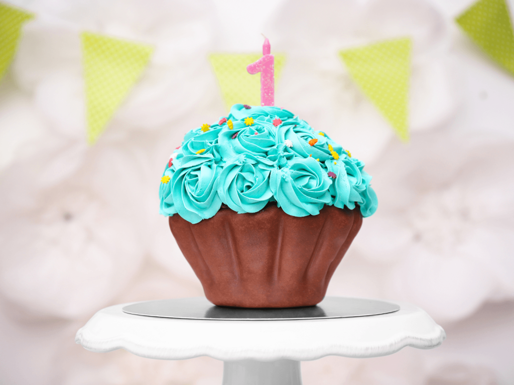 Cupcake-shaped first birthday cake with bright blue frosting, colorful sprinkles, and a pink number one candle on top.