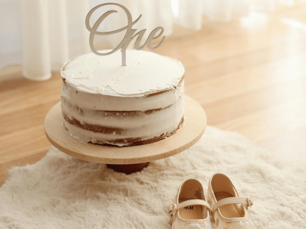 White first birthday cake topped with the word “One” beside a pair of tiny baby shoes.