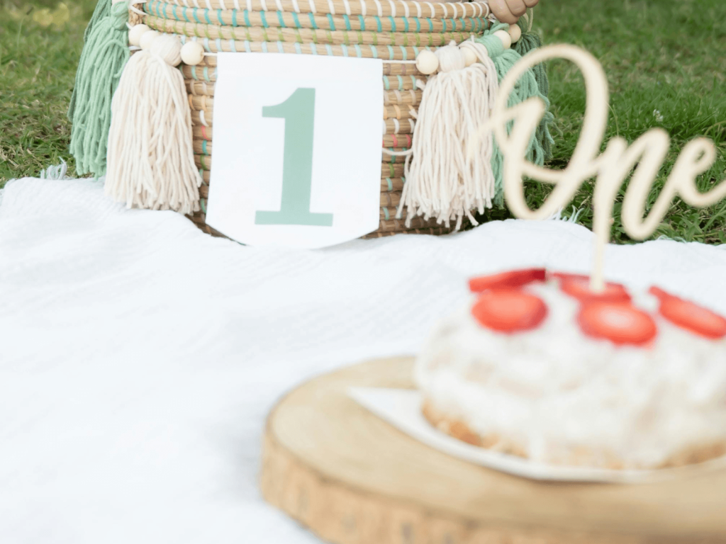 Outdoor first birthday cake smash setup with a number one sign, woven basket, and strawberry topped cake, perfect for 1st birthday wishes.