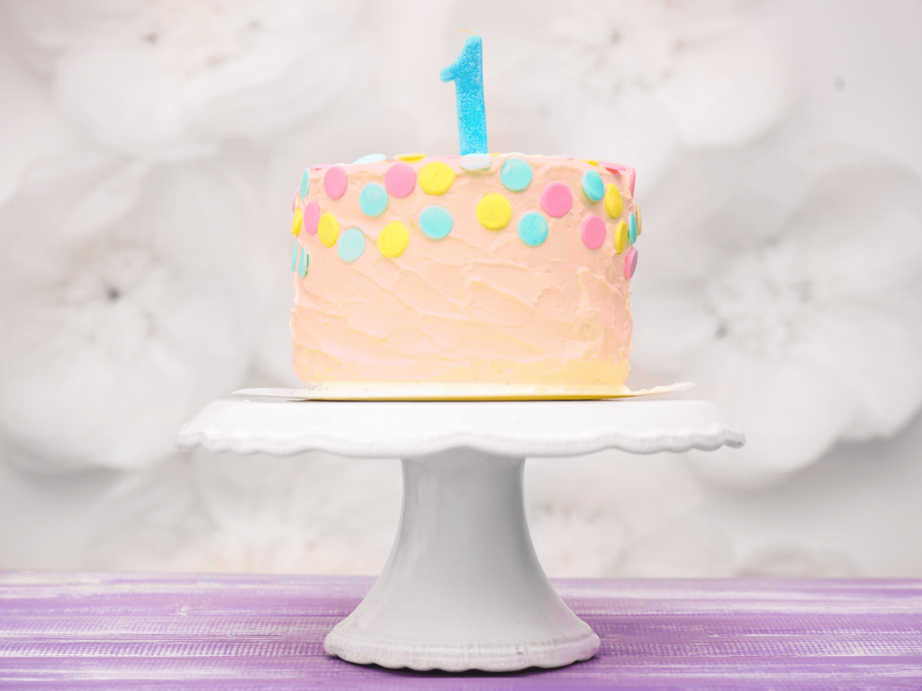 Pink first birthday cake on a white cake stand with pastel candy dots and a blue number one candle.