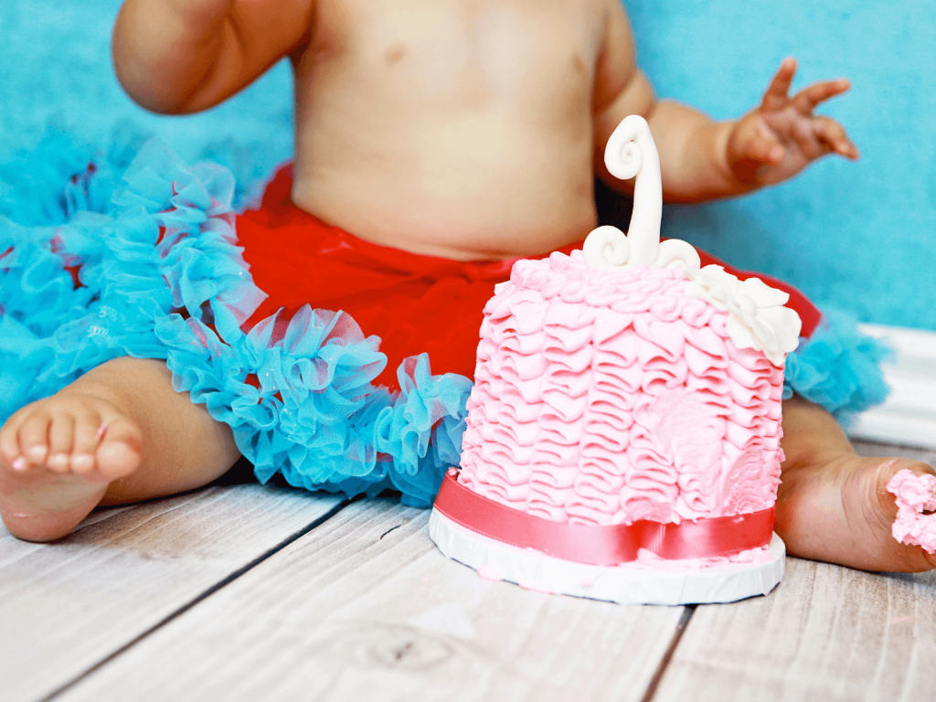 Baby sitting beside a pink first birthday cake with a white number one topper during a colorful cake smash photo session.