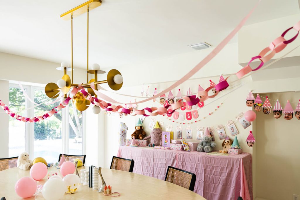 Dining room decorated with pink paper chains, balloons, stuffed animals, and baby photo banners for a first birthday party at home.