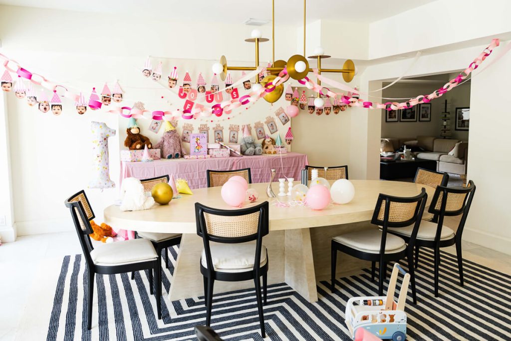 Dining room decorated with pink paper garlands, balloons, stuffed animals, and a first birthday photo banner for a baby’s first birthday party at home.