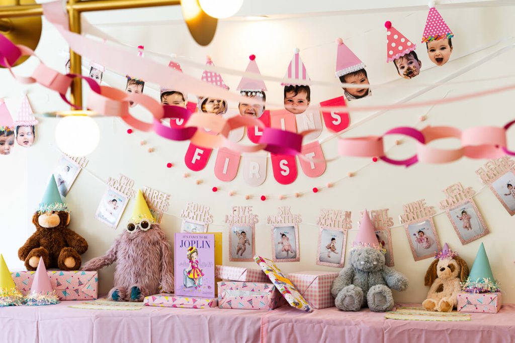 Pink first birthday party table with stuffed animals, wrapped gifts, paper chains, and monthly baby photo banners hanging on the wall.