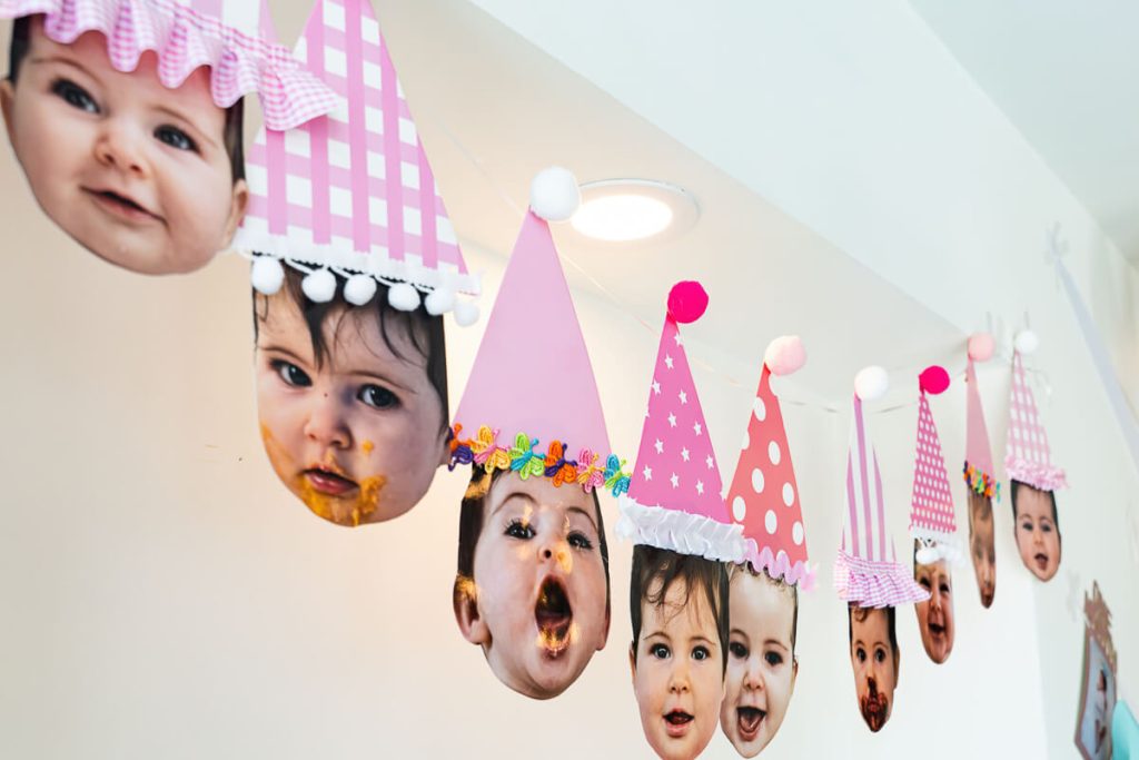 DIY first birthday photo banner with baby pictures wearing pink party hats strung across the wall as decoration.