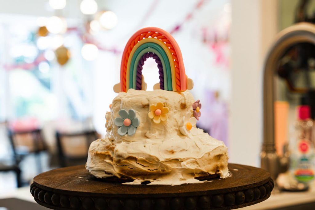 Homemade first birthday smash cake with white frosting, flower decorations, and a colorful rainbow topper on a cake stand.