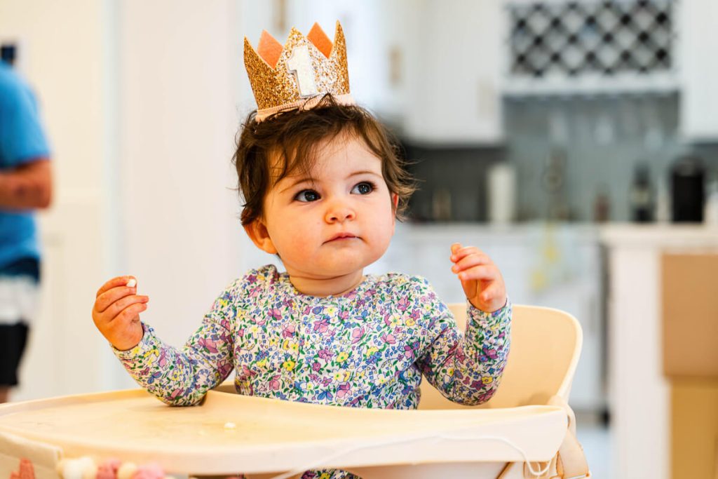 Baby wearing a gold number one crown sitting in a high chair during a first birthday party.