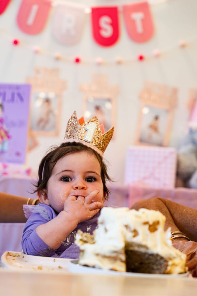 Close-up of a baby wearing a gold number one crown eating smash cake in front of pink first birthday decorations.