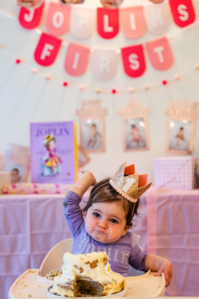 Baby wearing a gold number one crown sitting in a high chair with a partially eaten smash cake during a pink first birthday party.
