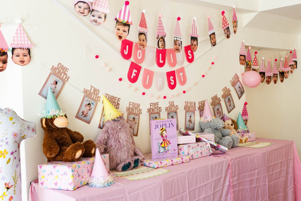 Pink first birthday party table decorated with stuffed animals, wrapped gifts, party hats, balloons, and monthly baby photo banners.