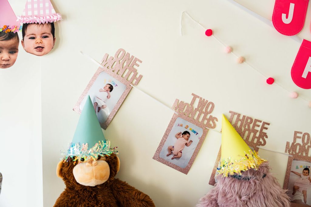 Monthly baby photo banner with one month, two months, and three months pictures displayed above stuffed animals wearing party hats for a first birthday party.