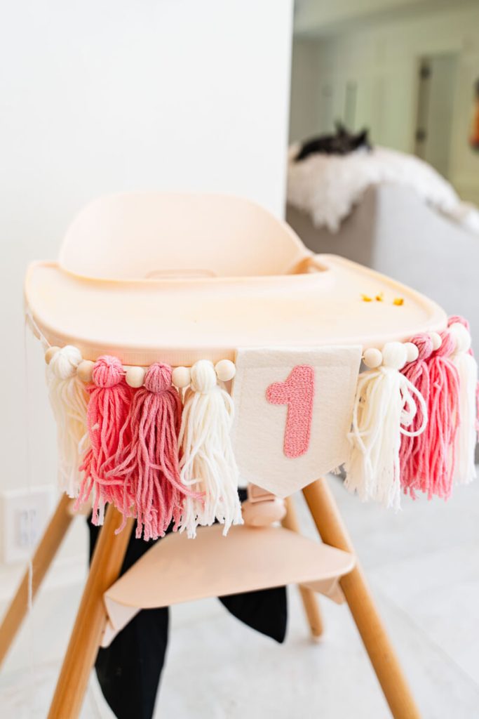 High chair decorated with a pink and white yarn banner and a number one sign for a first birthday smash cake setup.