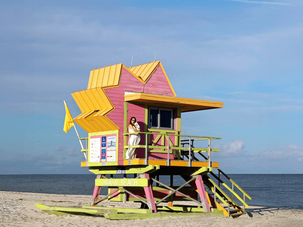 Mom holding her baby beside a colorful Miami Beach lifeguard tower, during a family outing with baby beach essentials.