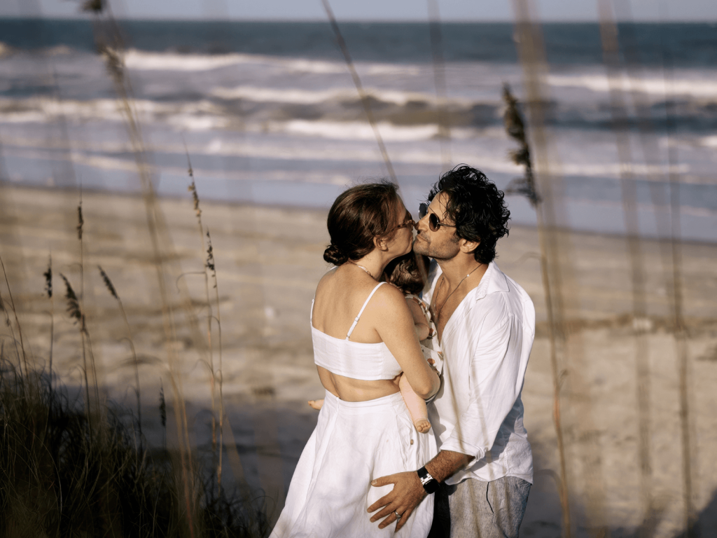 Parents in white clothing kiss while holding their baby on a sandy beach with ocean waves in the background.