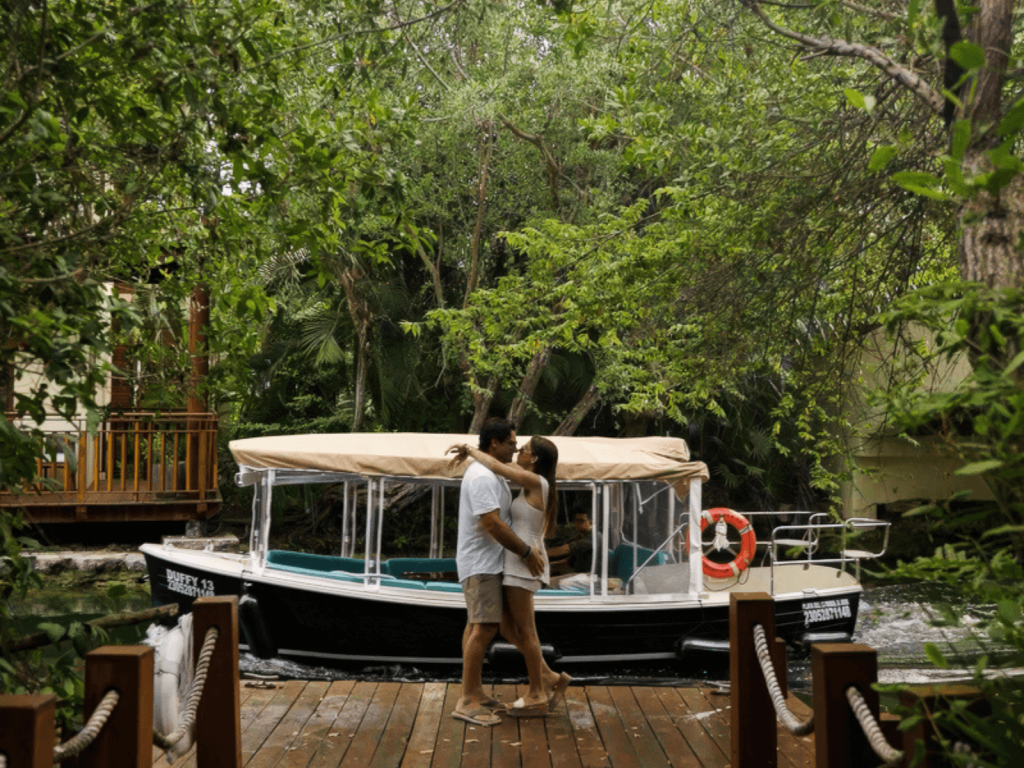 Couple embracing on a wooden dock beside a resort boat surrounded by lush mangroves and tropical greenery at Fairmont Mayakoba.