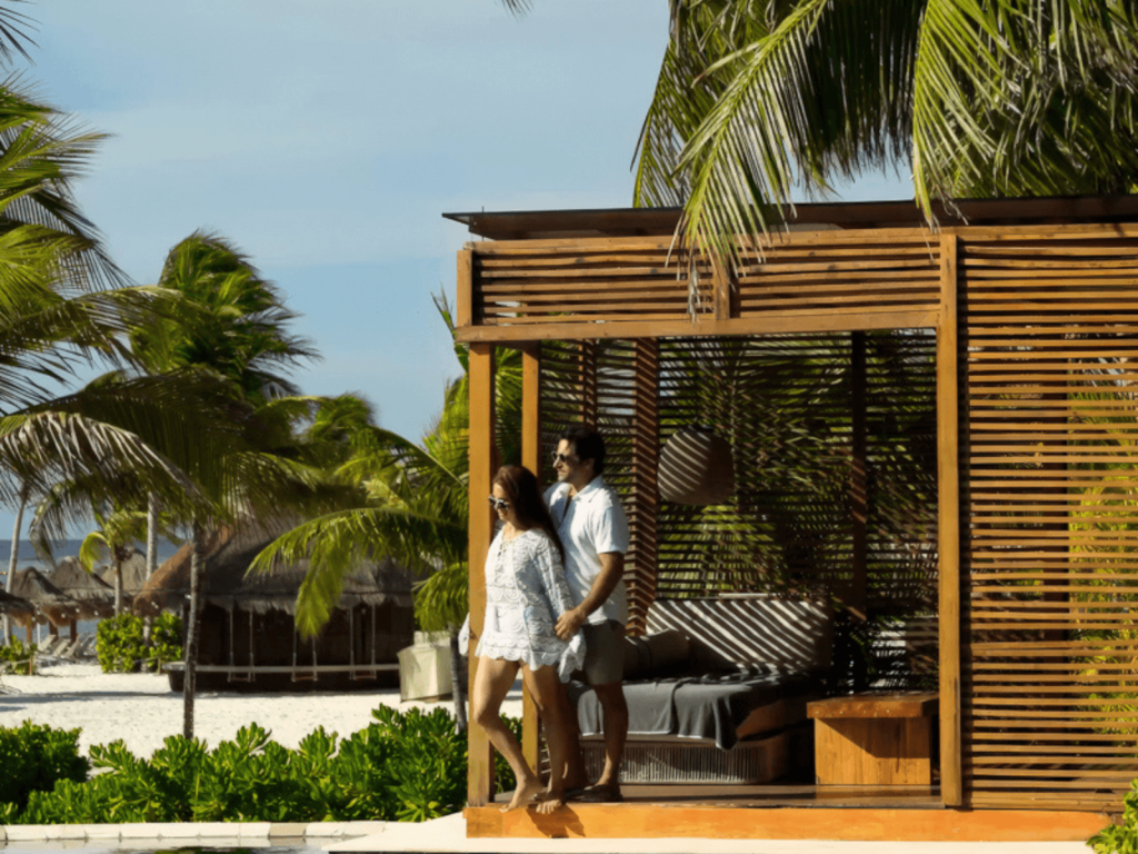 Couple standing beside a wooden beach cabana surrounded by palm trees with Fairmont Mayakoba beach in the background.