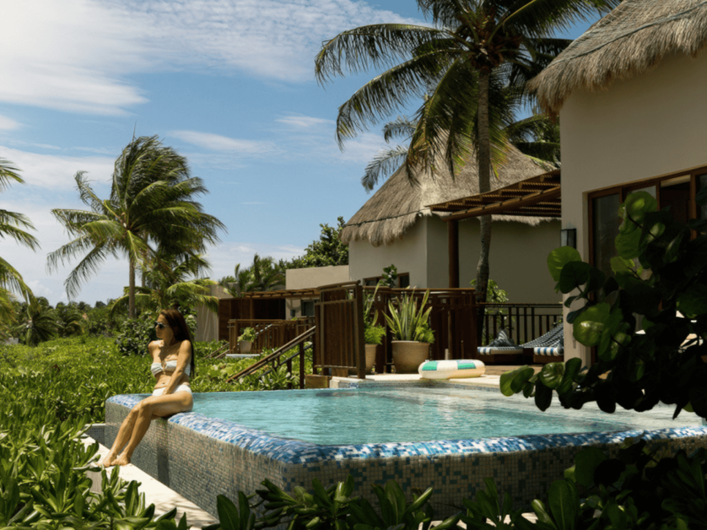 Woman sitting at the edge of a private pool beside a thatched roof casita surrounded by palm trees and tropical greenery at Fairmont Mayakoba.