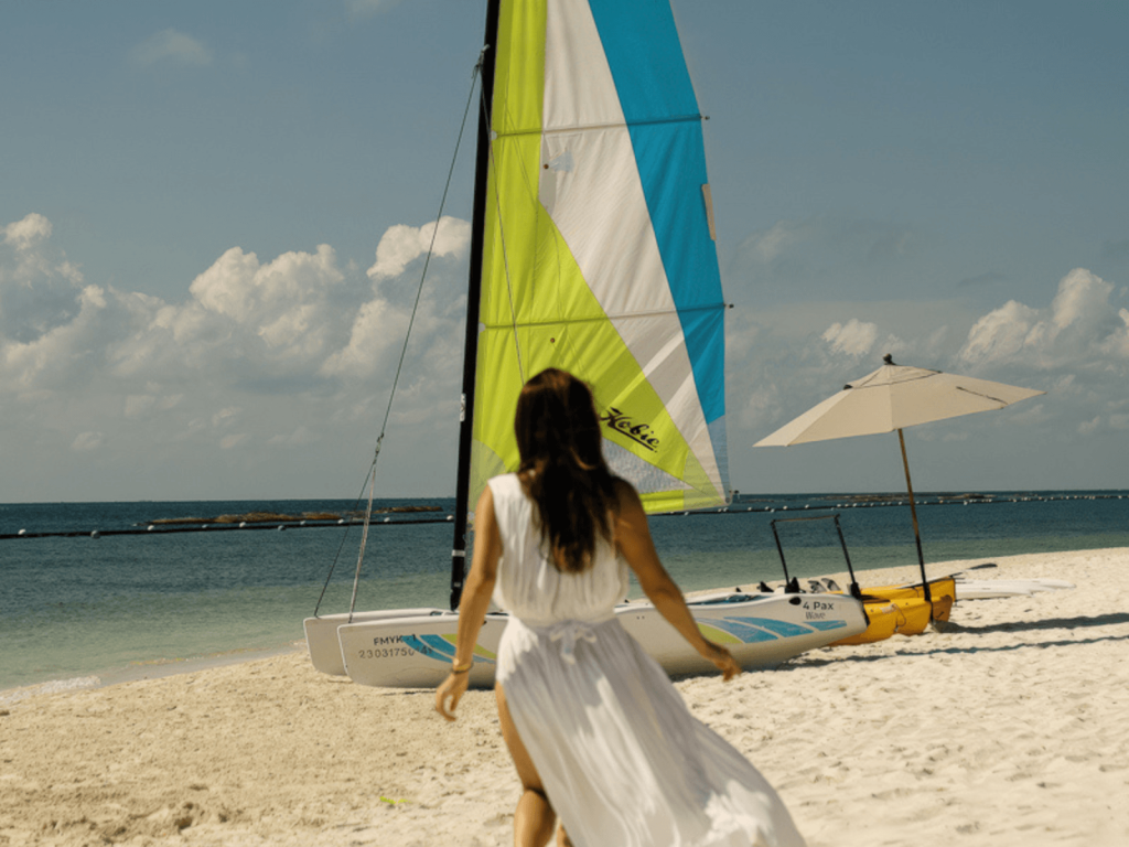 Woman in a white cover up walking toward a colorful sailboat on the sandy beach at Fairmont Mayakoba, Mexico.