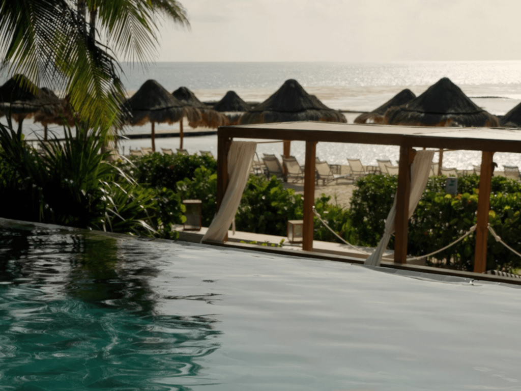 Infinity pool overlooking Fairmont Mayakoba beach with lounge chairs, palapas, palm trees, and the ocean in the background.