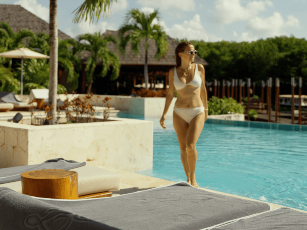 Woman in a white swimsuit walking through a turquoise pool surrounded by lounge chairs and palm trees at Fairmont Mayakoba.