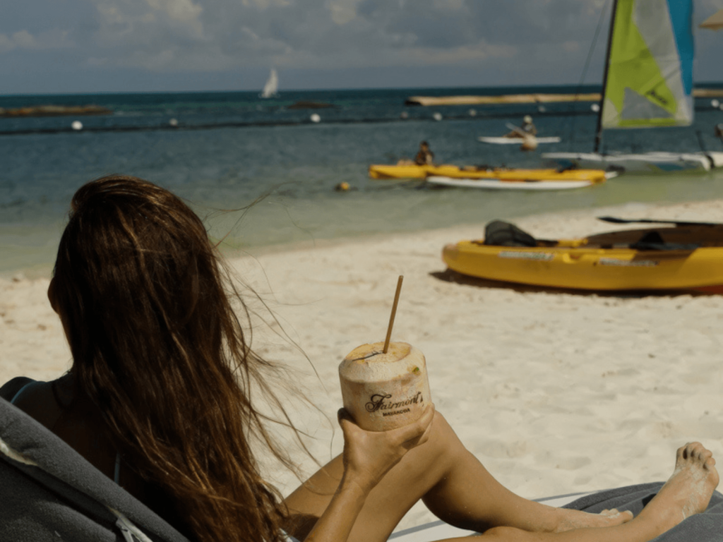 Woman relaxing on a beach lounger with a fresh coconut drink overlooking kayaks and sailboats at Fairmont Mayakoba beach.