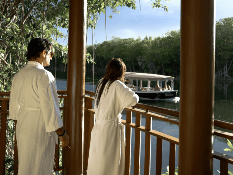 Couple in white robes standing on a wooden balcony overlooking a boat cruising through the mangrove lined waterways at Fairmont Mayakoba, Mexico, featured in a Fairmont Mayakoba review.
